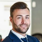 Caucasian businessman leader looking at camera in modern office with multi-ethnic businesspeople working at the background. Teamwork concept. Young man with beard wearing blue suit.